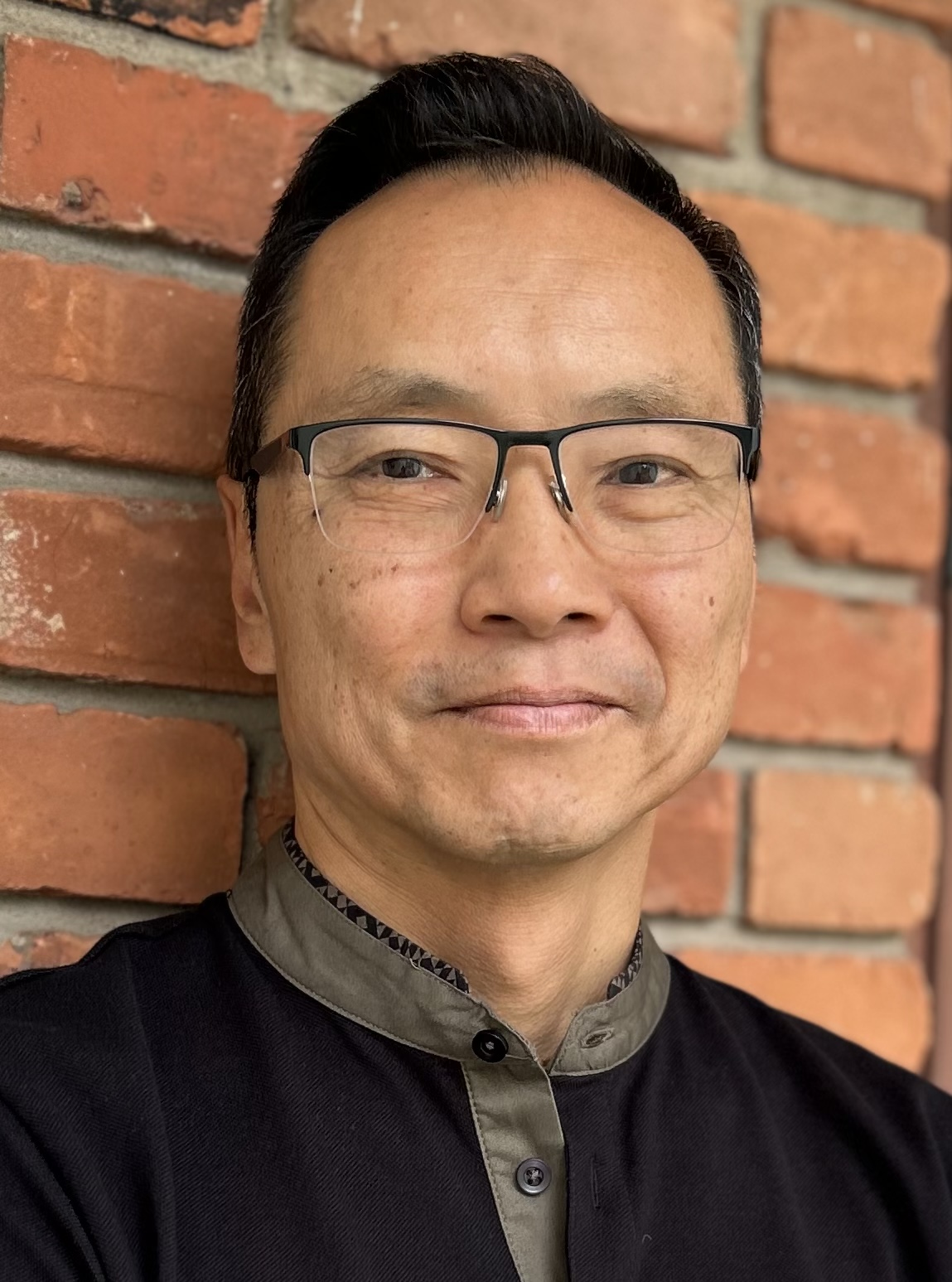 Author photo of Wayne Ng. A head and shoulders shot of Wayne Ng in a black shirt with a grey collar against a red brick background.